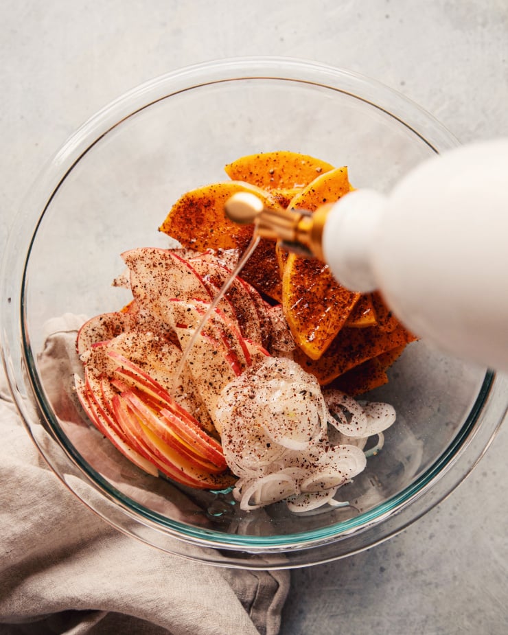 An overhead shot of a galette filling: roasted butternut squash, sliced apples, and shallots. Olive oil is being drizzled in.