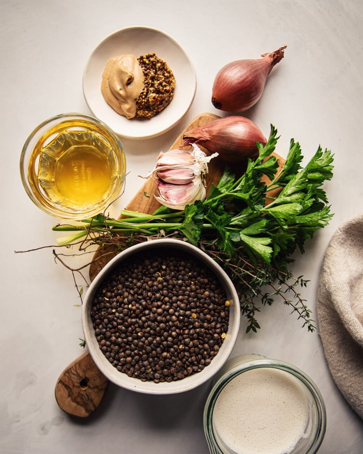 An overhead shot of the ingredients used for a stewed lentil dish. There's shallots, garlic, grainy and Dijon mustard, white wine, parsley, thyme, French lentils, and a cream made out of cashews.