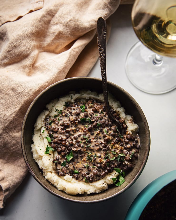 An overhead shot of white wine lentils with mustard and thyme in an olive green bowl. The beige, stewed lentils are served over mashed cauliflower. The bowl is served alongside a pale yellow napkin and a glass of white wine, all on a white background. Part of a roundup of vegan soup recipes.