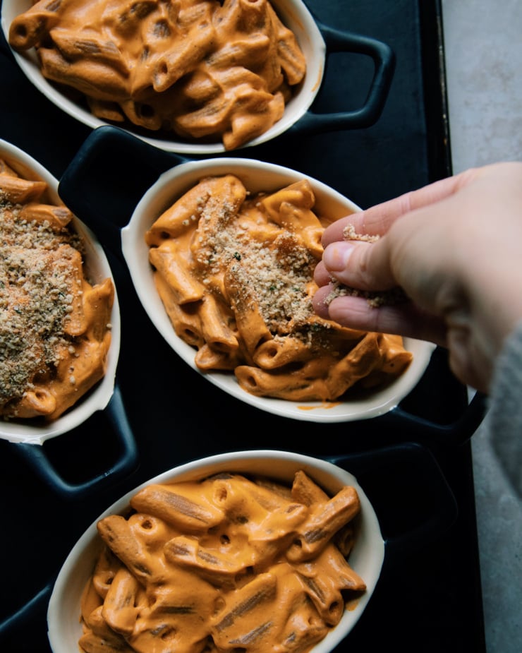An overhead shot of a hand sprinkling cracker topping onto little black ramekins of creamy red pepper pasta.