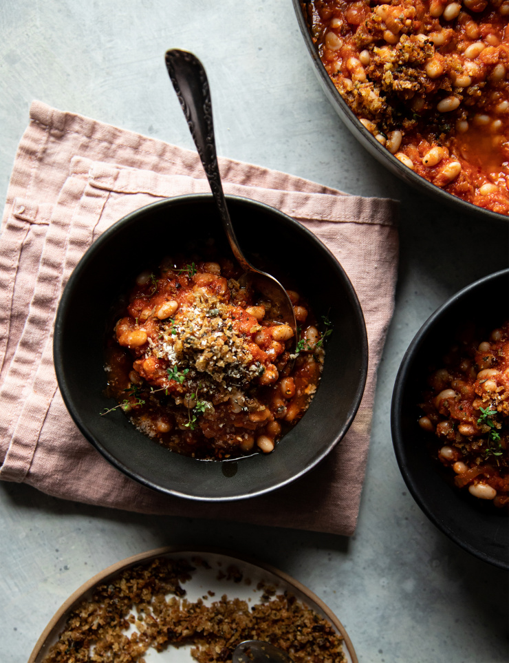 Overhead shot of a single serving of jammy sun dried tomato white beans. The white beans are plated in a matte back bowl on top of a pink napkin.