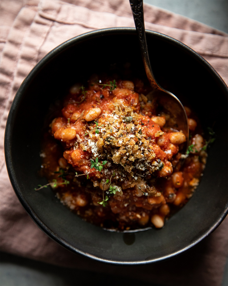 Overhead, up close shot of a single serving of jammy sun dried tomato white beans. The white beans are plated in a matte back bowl on top of a pink napkin.