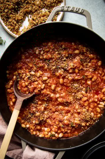 An overhead shot of jammy sun dried tomato white beans in a beige braiser-style pot. To the side there is a plate of crispy caper breadcrumbs and a pink napkin.