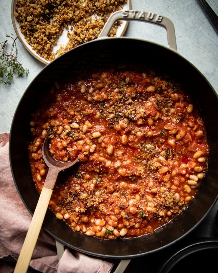 An overhead shot of jammy sun dried tomato white beans in a beige braiser-style pot. To the side there is a plate of crispy caper breadcrumbs and a pink napkin.