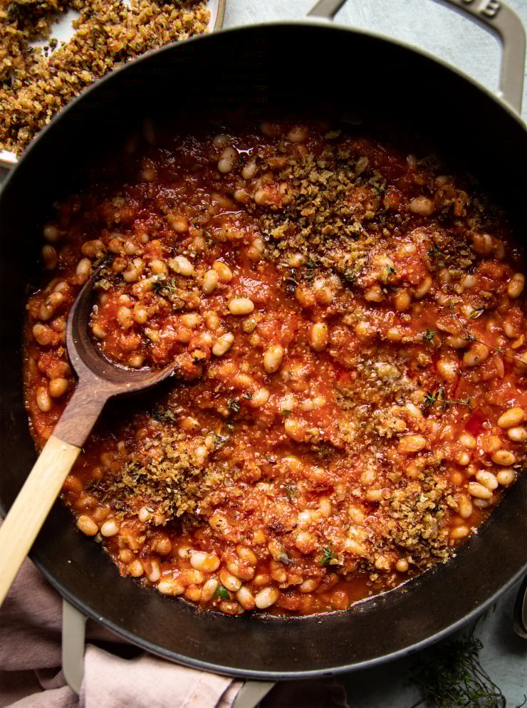 An overhead shot of jammy sun dried tomato white beans in a beige braiser-style pot. The white beans are garnished with crispy cakes breadcrumbs and thyme leaves. There is a wooden spoon sticking out of the pot.