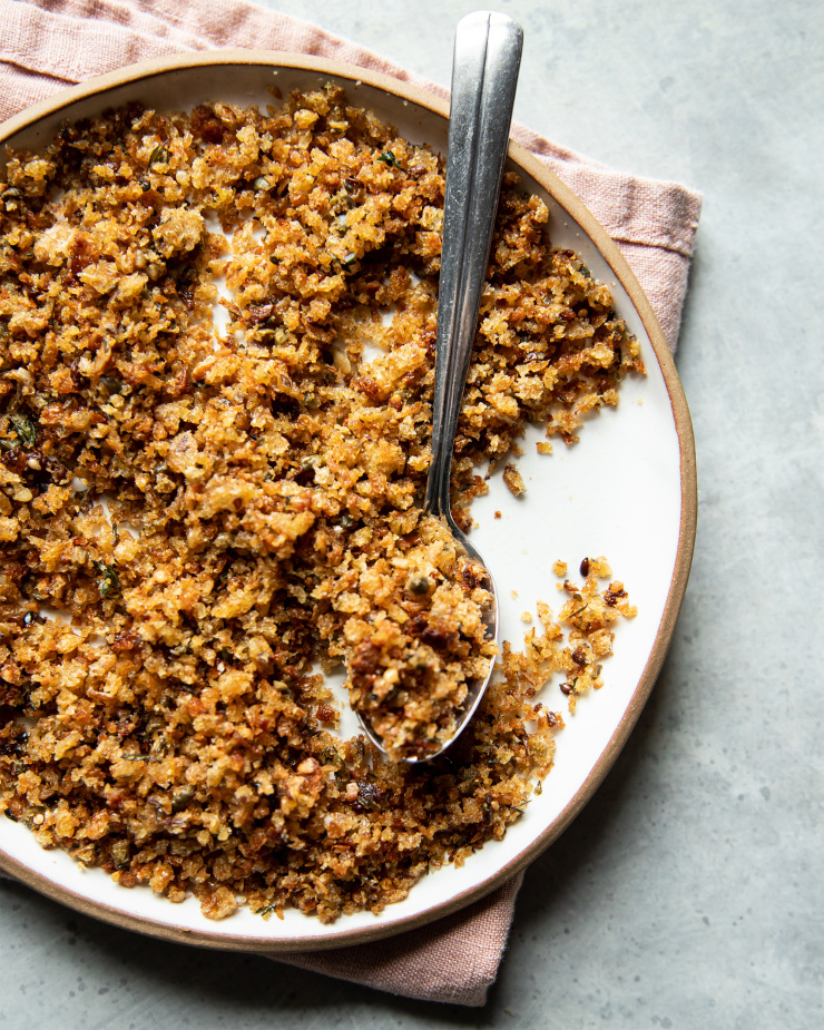 An overhead shot of crispy caper breadcrumbs in a white, lipped plate. There is a spoon on the plate.