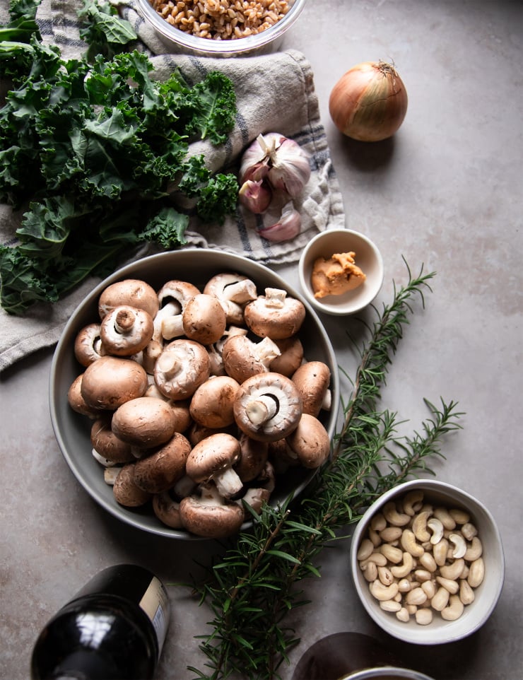 An overhead shot of ingredients on a grey stone background. There is: kale, an onion, a head of garlic, cooked farro in a bowl, a bowl of Cremini mushrooms, sprigs of rosemary, and cashews.