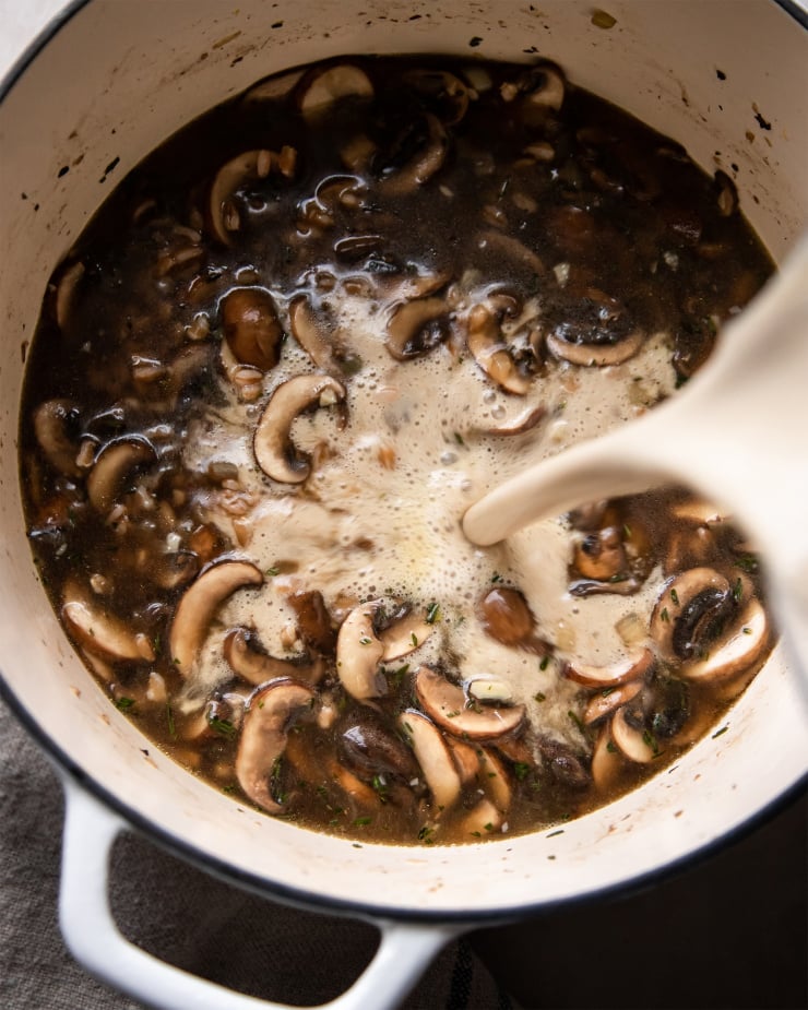 An overhead shot of bright white cashew cream being poured into a pot with deep brown mushroom stew.