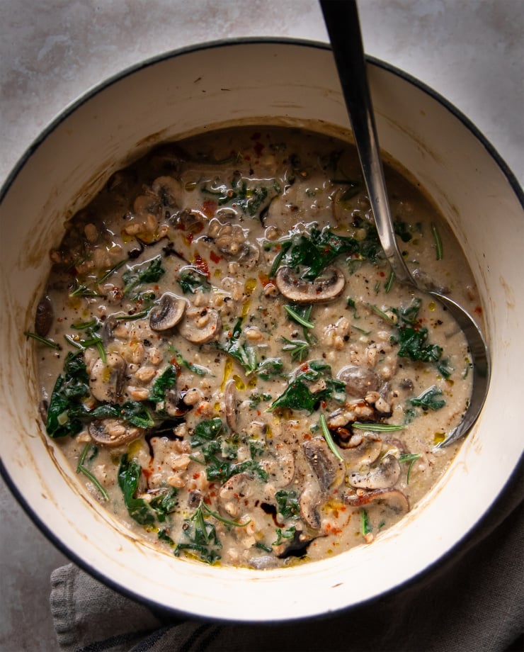Overhead shot of creamy vegan mushroom stew with farro and kale. The stew is a golden beige colour and is photographed in a white Dutch oven pot on a grey background.