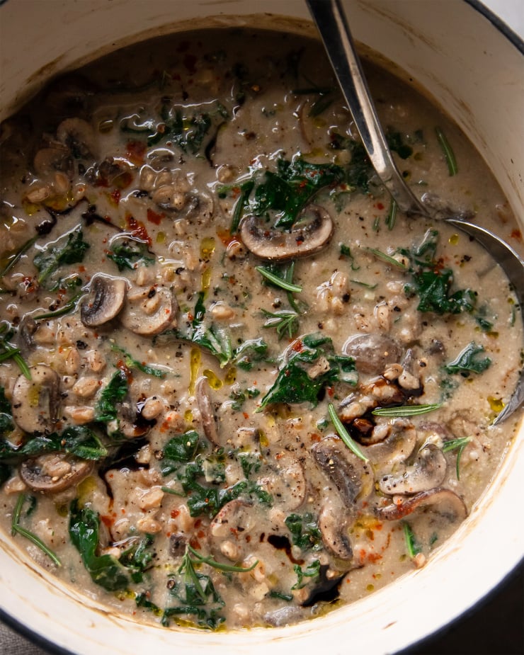 AN up close, overhead shot of creamy vegan mushroom stew with kale and farro. The soup is a golden beige colour and is inside a white Dutch oven pot.