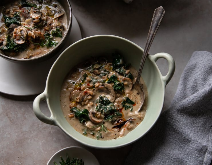An overhead shot of creamy vegan mushroom stew with faro and kale in a light green bowl with handles on the side. The bowl is set on a grey stone background in low lighting.