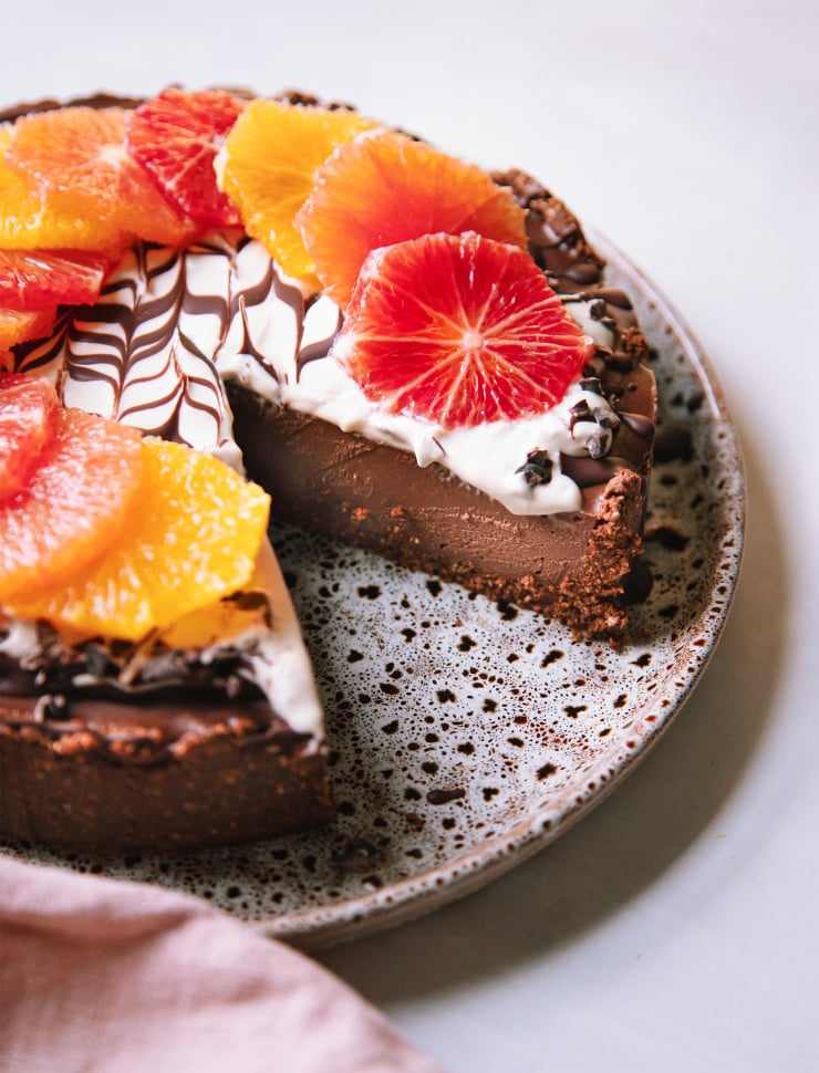 A side angle shot of a vegan chocolate orange cheesecake with a slice removed, showing the chocolaty truffle-like texture of the cake. The top of the cheesecake is decorated with different coloured orange slices and a bright white cashew cream marbled with melted chocolate.