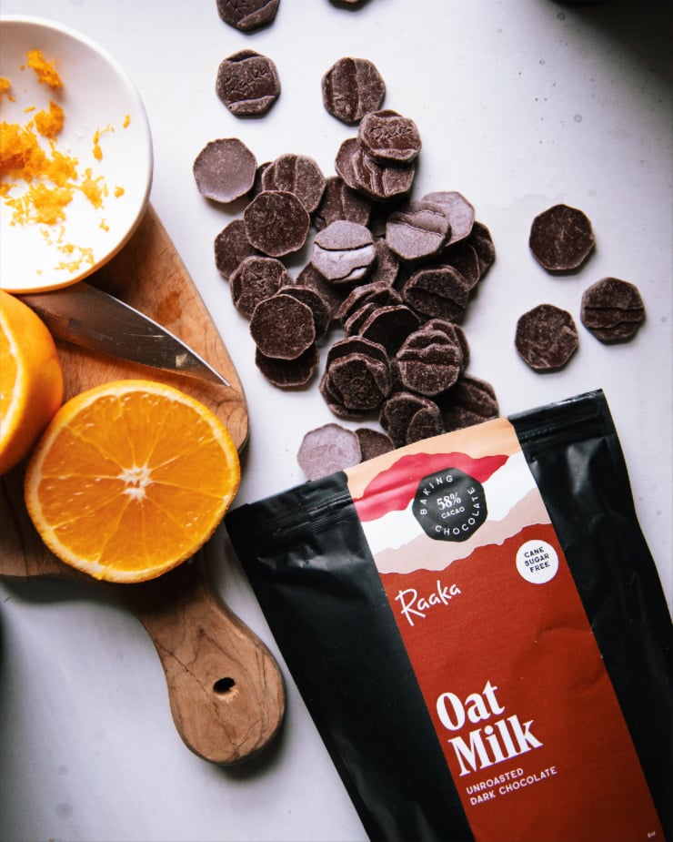 An overhead shot of a bag of Raaka Oat Milk Baking Chocolate spilling out onto a white background. There is a sliced orange and a small white bowl of orange zest nearby.