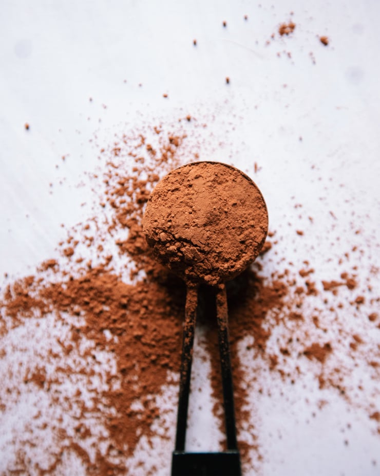 An overhead shot of a black measuring spoon filled with cacao powder on a white background. The cacao is spilled out all around the spoon.