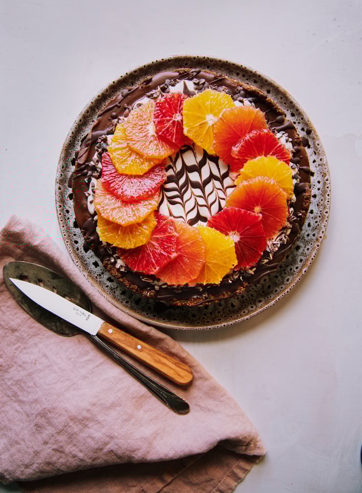 An overhead shot of a vegan chocolate orange cheesecake on a white background. The top of the cheesecake has a wreath or different coloured orange slices on top of a white cashew cream and melted chocolate marble design. To the side is a pink linen napkin, a paring knife and a vintage silver serving utensil.