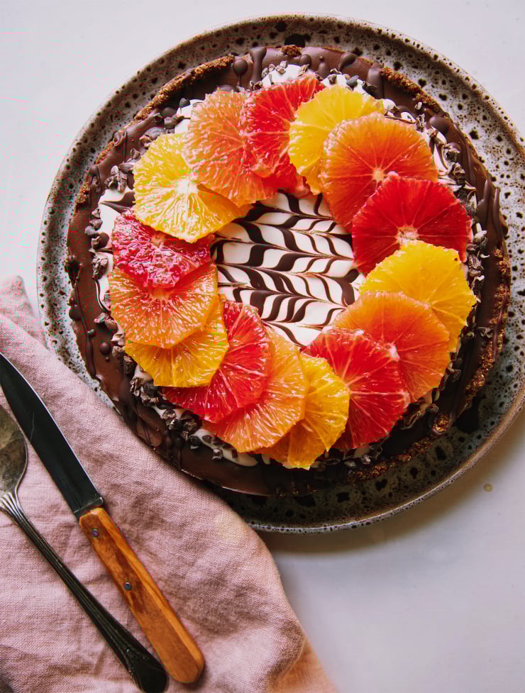 An overhead shot of a vegan chocolate orange cheesecake on a white background. The top of the cheesecake has a wreath or different coloured orange slices on top of a white cashew cream and melted chocolate marble design. To the side is a pink linen napkin, a paring knife and a vintage silver serving utensil.
