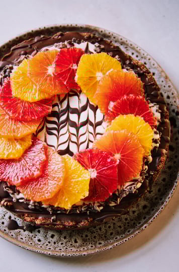 An overhead shot of a vegan chocolate orange cheesecake on a white background. The top of the cheesecake has a wreath or different coloured orange slices on top of a white cashew cream and melted chocolate marble design. To the side is a pink linen napkin, a paring knife and a vintage silver serving utensil.