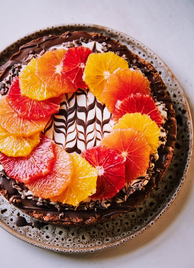 An overhead shot of a vegan chocolate orange cheesecake on a white background. The top of the cheesecake has a wreath or different coloured orange slices on top of a white cashew cream and melted chocolate marble design. To the side is a pink linen napkin, a paring knife and a vintage silver serving utensil.