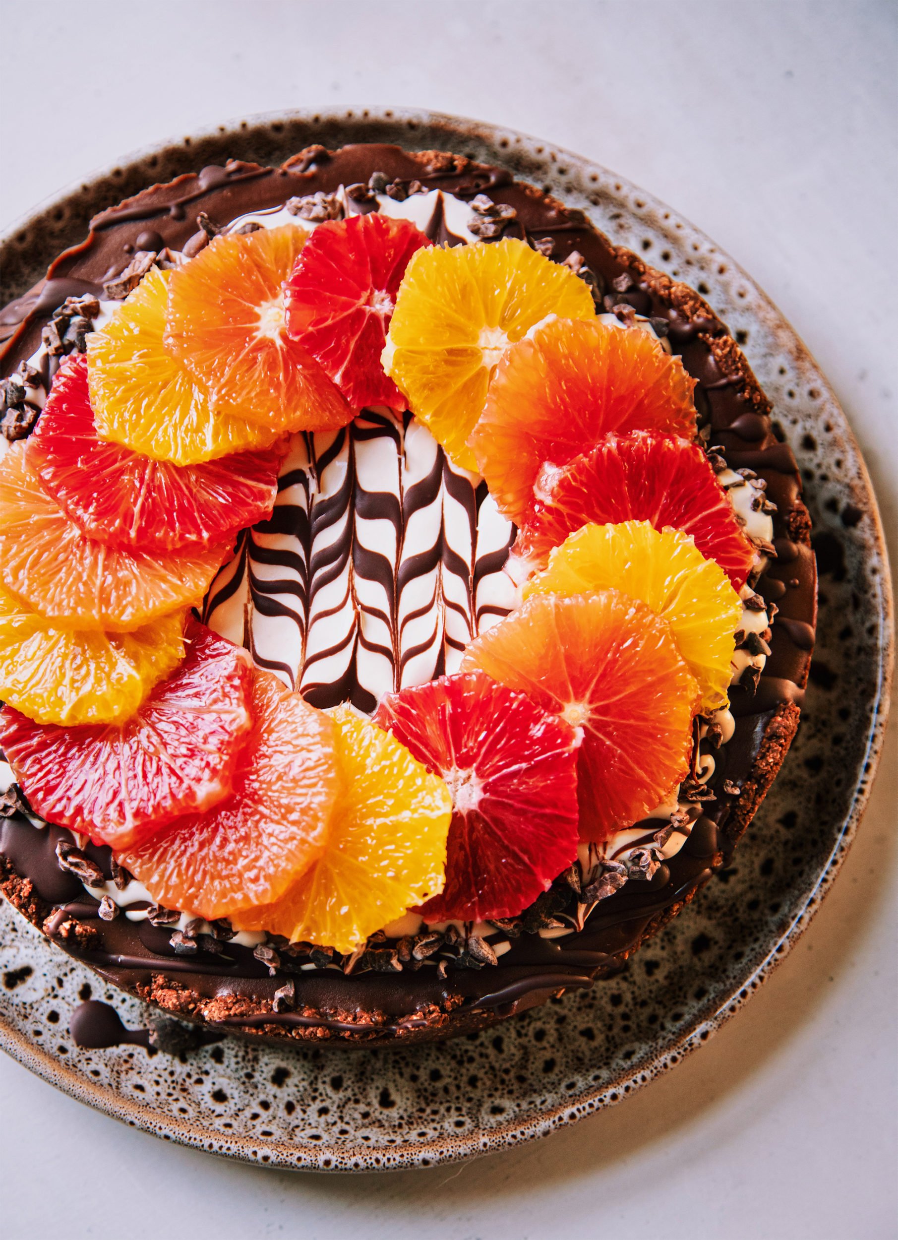 An overhead shot of a vegan chocolate orange cheesecake on a white background. The top of the cheesecake has a wreath or different coloured orange slices on top of a white cashew cream and melted chocolate marble design. To the side is a pink linen napkin, a paring knife and a vintage silver serving utensil.