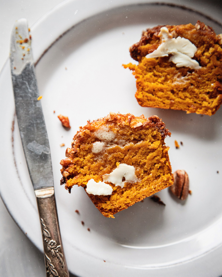 An up close, overhead shot of a split vegan sweet potato muffin on a white plate with a little bit of vegan butter smeared onto the cut side. There is a butter knife perched on the plate as well.