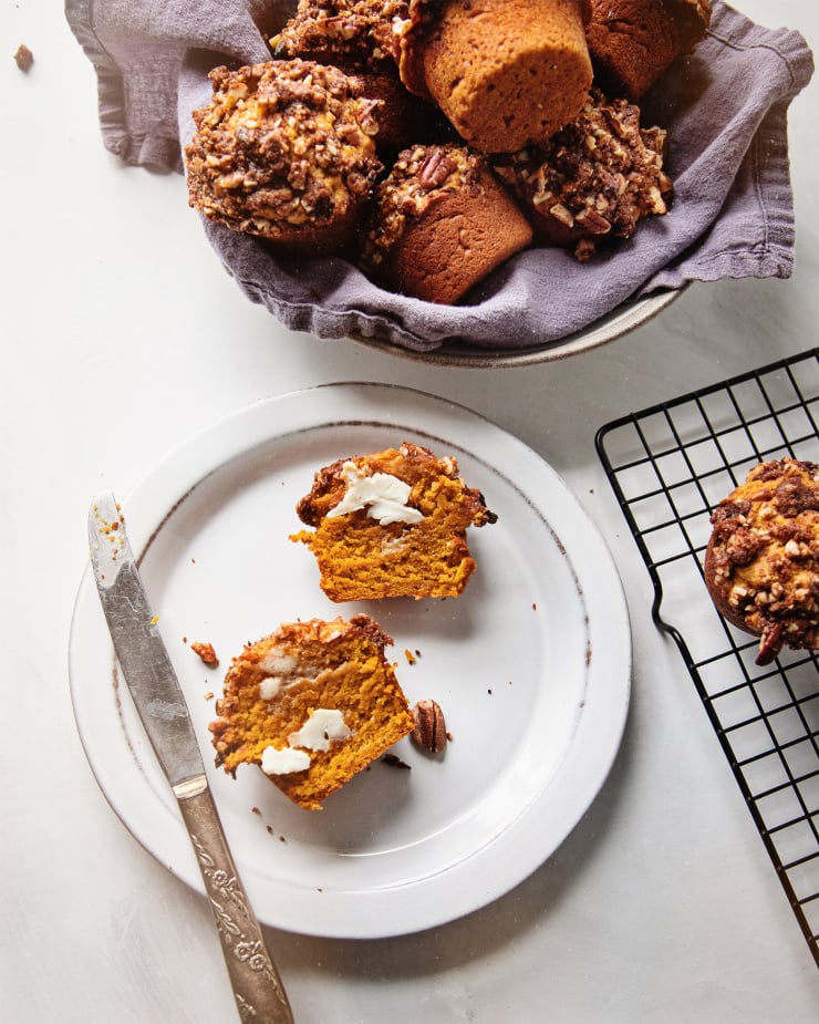 An overhead shot of vegan sweet potato muffins in a bowl lined with a navy linen napkin. To the side is a white plate with a split open muffin and a butter knife.