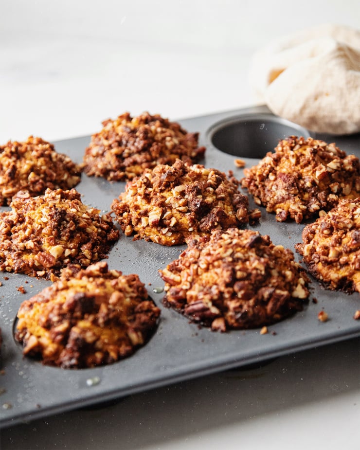 A side angle shot of baked sweet potato muffins in a muffin tin. The tops are domed, the muffins are slightly orange-hued, and the pecan streusel is golden brown.