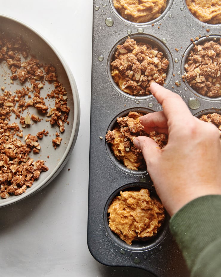 An overhead shot of a hand applying pecan streusel to sweet potato muffin batter in a muffin tin.