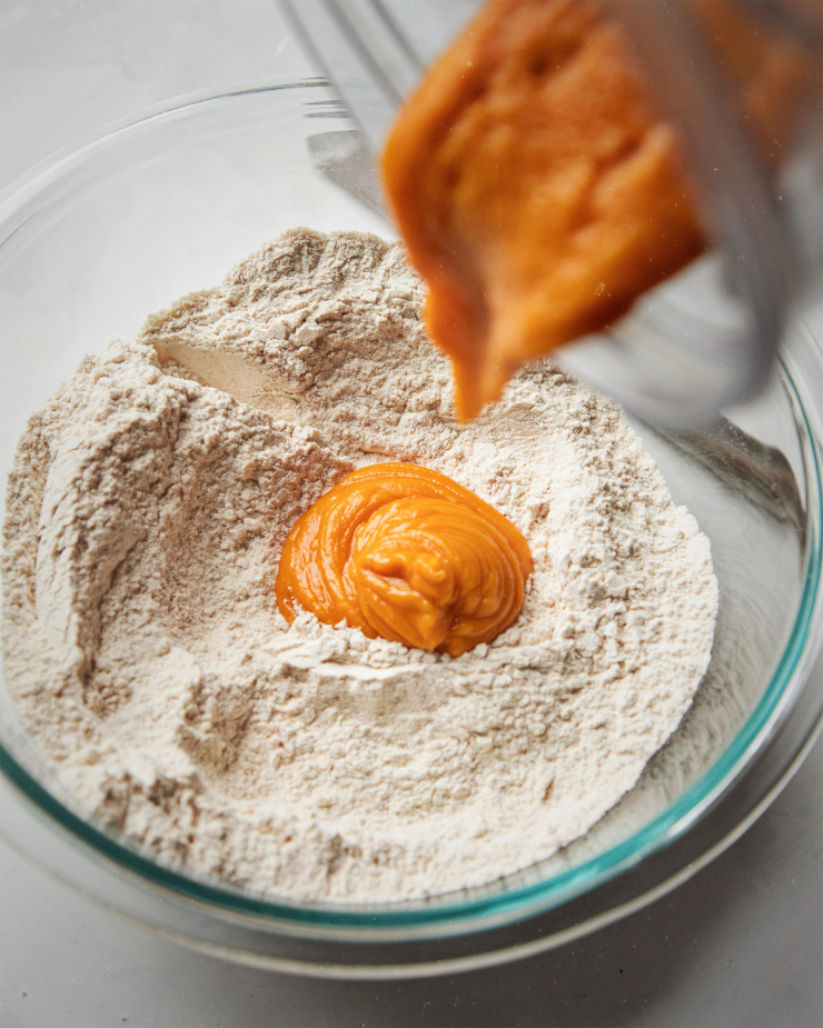 A 3/4 angle shot of sweet potato purée being poured into a glass bowl with dry ingredients for muffins. The purée is bright orange.