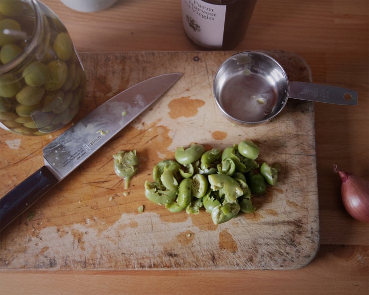 An overhead shot of Castelvetrano olives that. Have been crushed on a worn wood cutting board. A knife and the jar of olives are to the side.