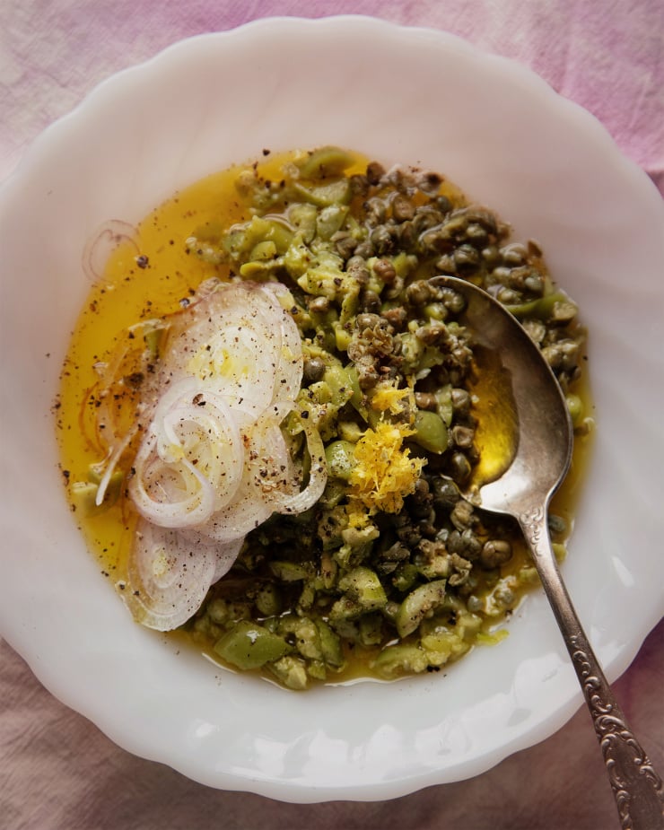 An overhead shot of a chunky olive, caper, and shallot dressing before it is whisked together. The ingredients are in a shallow, white milk glass bowl.