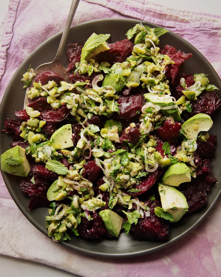 An overhead shot of a crushed beets salad with chunky olive dressing, mint, and avocado. The salad is on a matte black plate on top of a pink and purple tie-dyed linen.