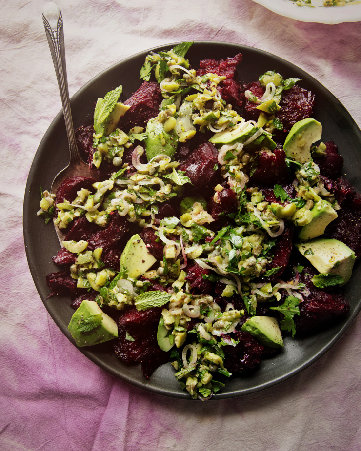 An overhead shot of a crushed beets salad with chunky olive dressing, mint, and avocado. The salad is on a matte black plate on top of a pink and purple tie-dyed linen.