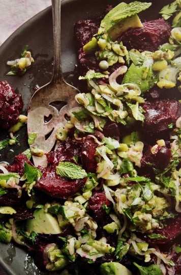 An up close, overhead shot of a crushed beets salad with chunky olive dressing, mint, and avocado. The salad is on a matte black plate. A vintage silver serving utensil has removed some of the salad.