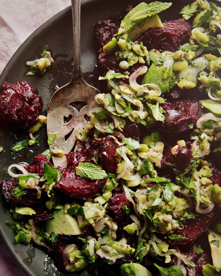 An up close, overhead shot of a crushed beets salad with chunky olive dressing, mint, and avocado. The salad is on a matte black plate. A vintage silver serving utensil has removed some of the salad.