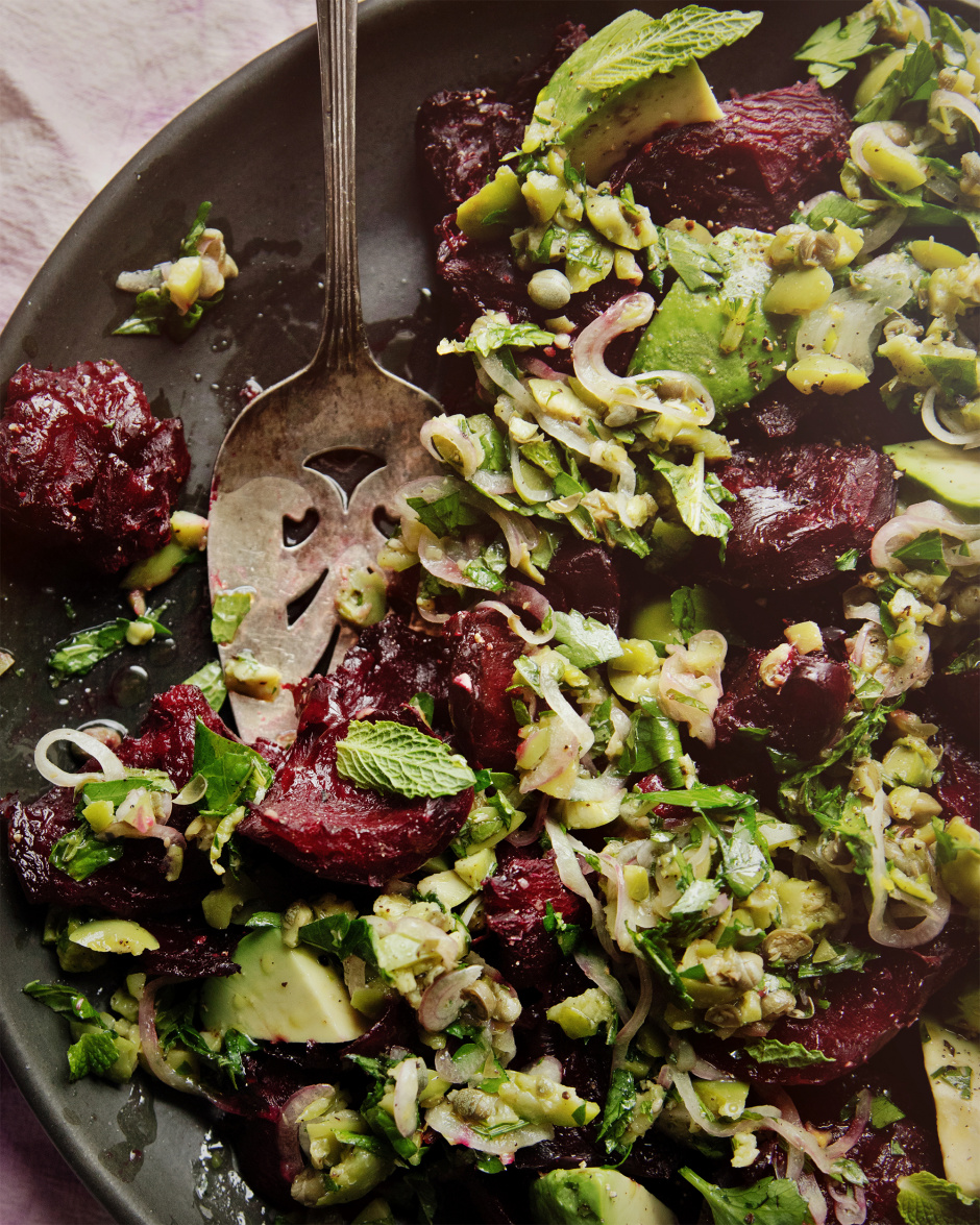 An up close, overhead shot of a crushed beets salad with chunky olive dressing, mint, and avocado. The salad is on a matte black plate. A vintage silver serving utensil has removed some of the salad.