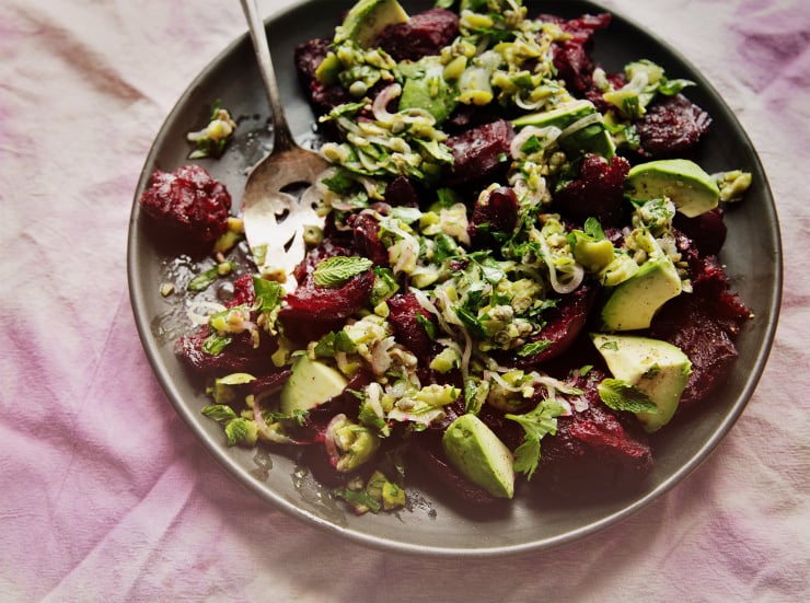 An overhead, slightly angled shot of a crushed beets salad with chunky olive dressing, mint, and avocado. The salad is on a matte black plate on top of a pink and purple tie-dyed linen.