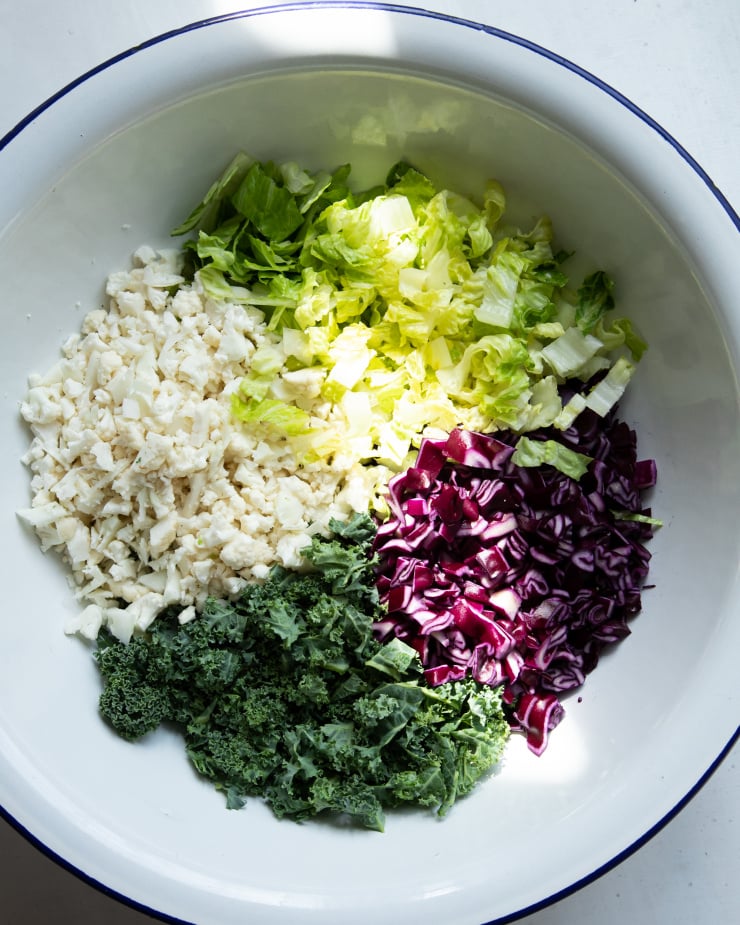 Image shows chopped vegetables in a large white bowl, before mixing into a salad with dressing.