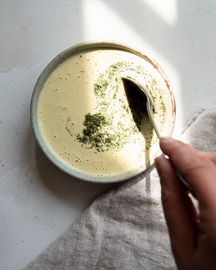 Image shows a hand stirring dried dill into a bowl of creamy dressing.