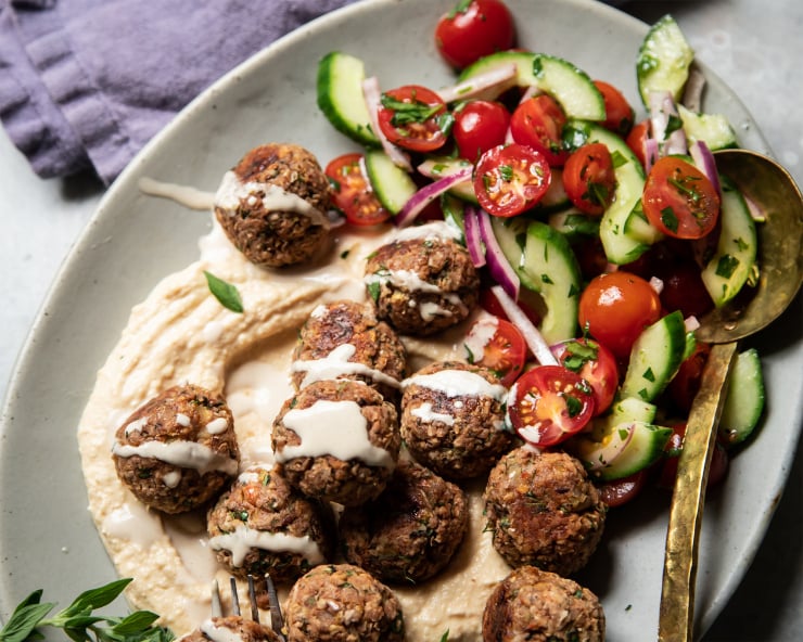 An overhead shot of meatballs on a platter with hummus, tahini sauce, and a cucumber tomato salad.