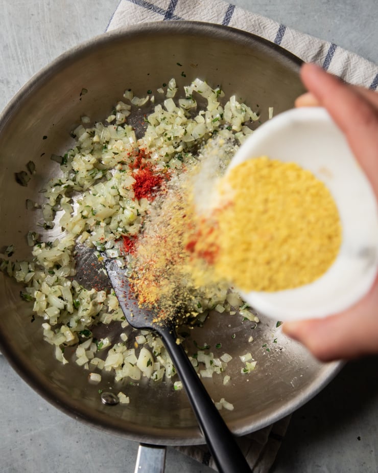 An overhead shot of a hand sprinkling spices into a sauté pan filled with onions and herbs.