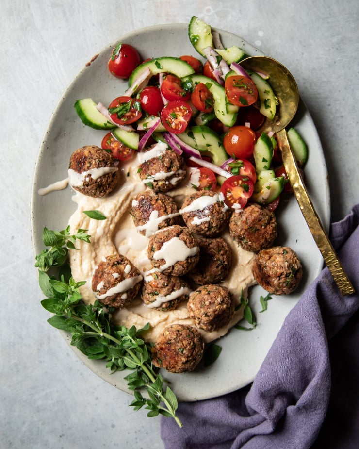 An overhead shot of vegan lemony oregano meatballs on a platter with hummus, tahini sauce, and a cucumber tomato salad.