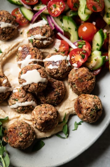 An overhead shot of vegan lemony oregano meatballs on a platter with hummus, tahini sauce, and a cucumber tomato salad.