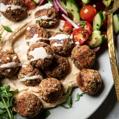 An overhead shot of vegan lemony oregano meatballs on a platter with hummus, tahini sauce, and a cucumber tomato salad.