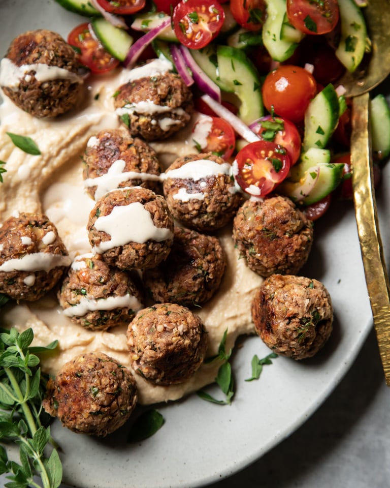 An overhead shot of vegan lemony oregano meatballs on a platter with hummus, tahini sauce, and a cucumber tomato salad.