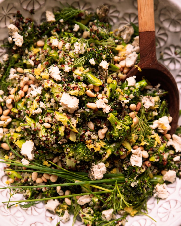 Image shows a green love quinoa salad in a patterned white bowl. The salad is garnished with whole blades of chives and bits of soft vegan cheese.