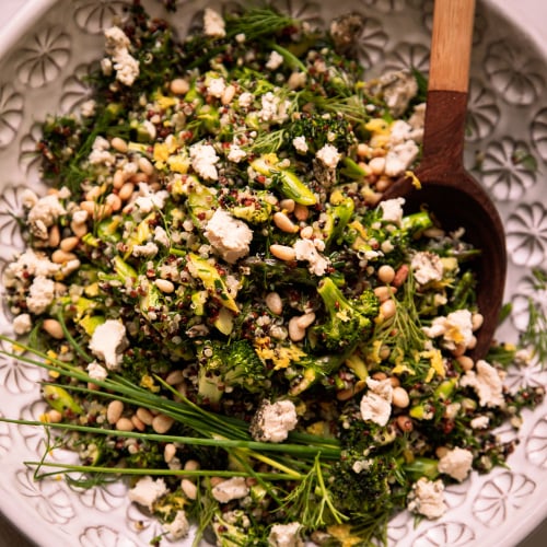 Image shows a green love quinoa salad in a patterned white bowl. The salad is garnished with whole blades of chives and bits of soft vegan cheese.