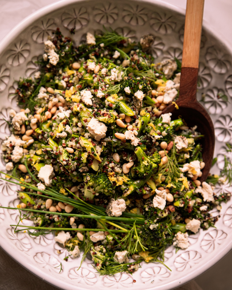 Image shows a green love quinoa salad in a patterned white bowl. The salad is garnished with whole blades of chives and bits of soft vegan cheese.