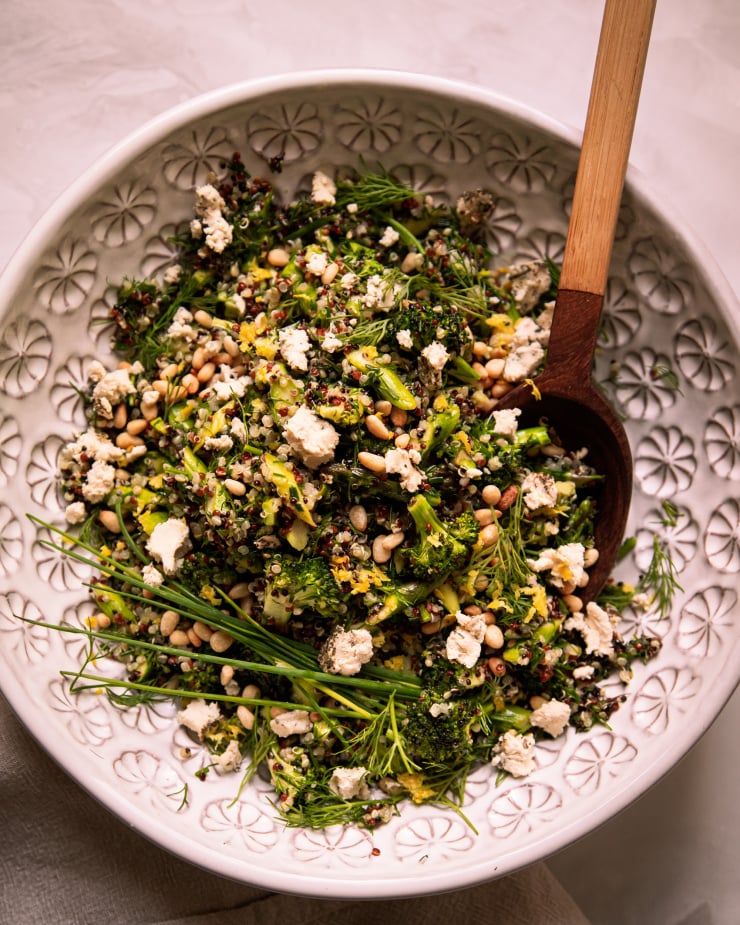 Image shows a green love quinoa salad in a patterned white bowl. The salad is garnished with whole blades of chives and bits of soft vegan cheese.