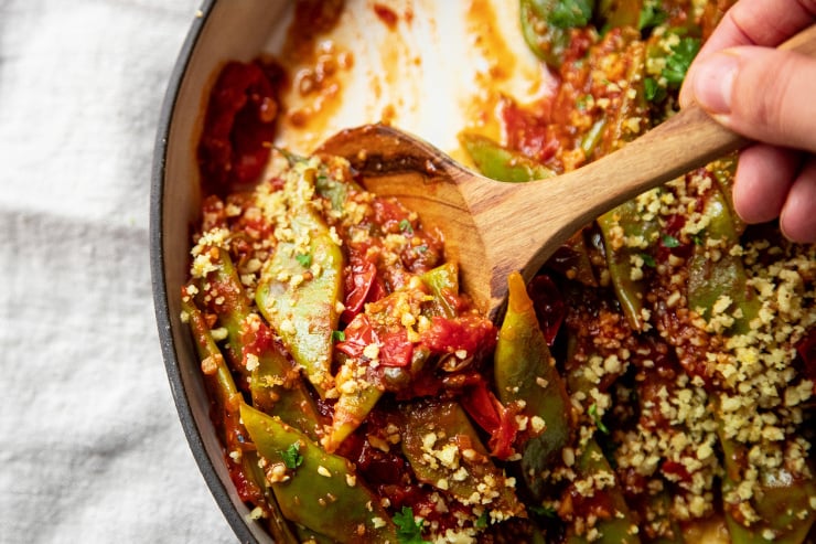 A hand is stirring some saucy green beans. An up close and overhead shot.