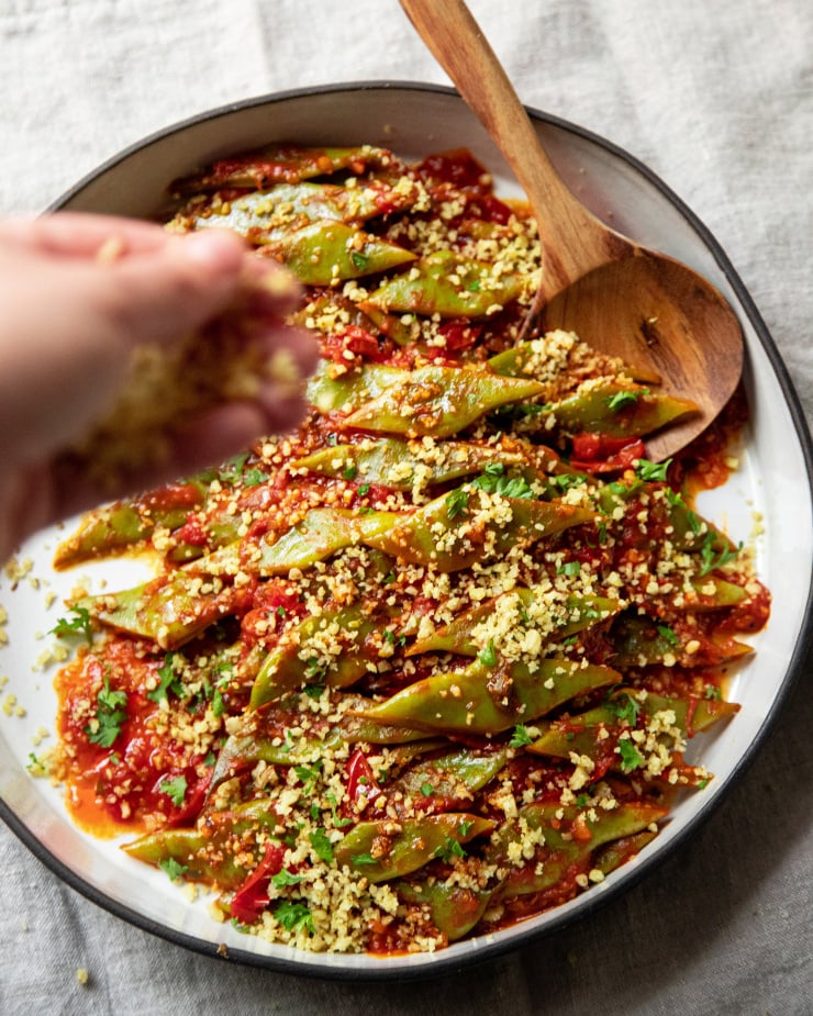 A hand is sprinkling ground walnuts on a Romano bean dish.